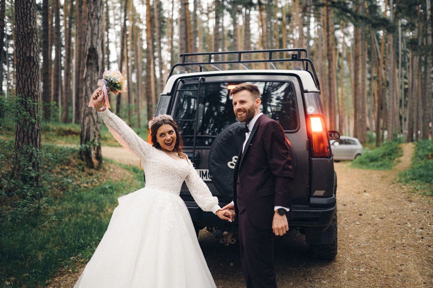 Joyful newlyweds celebrating near their off-road vehicle in pine forest.