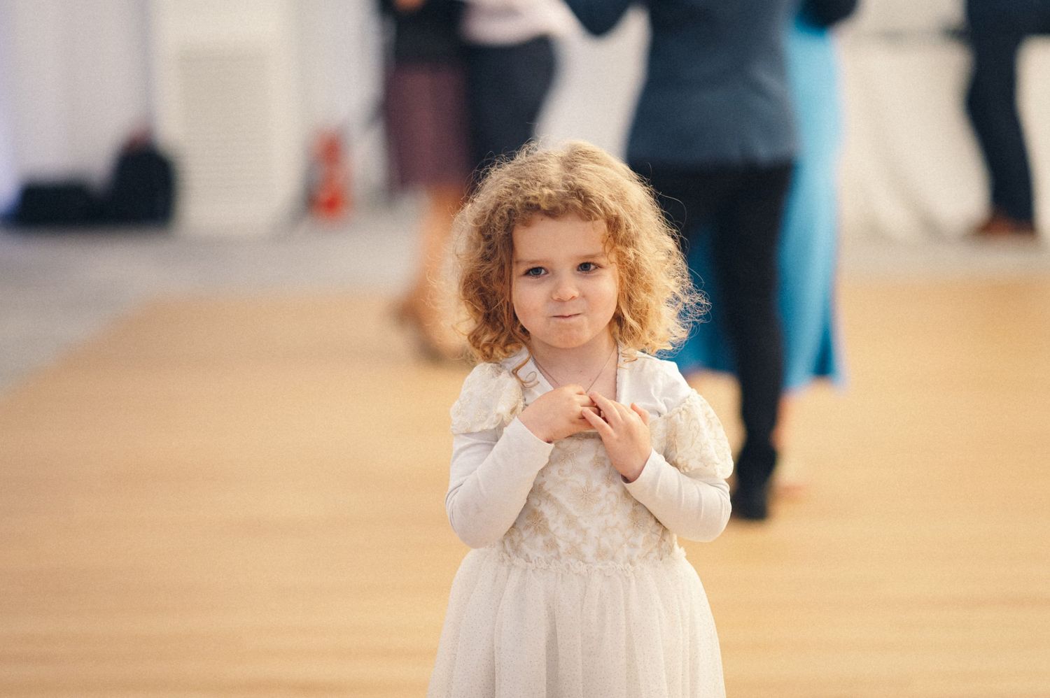 Young girl in white dress stands shyly on wooden dance floor.