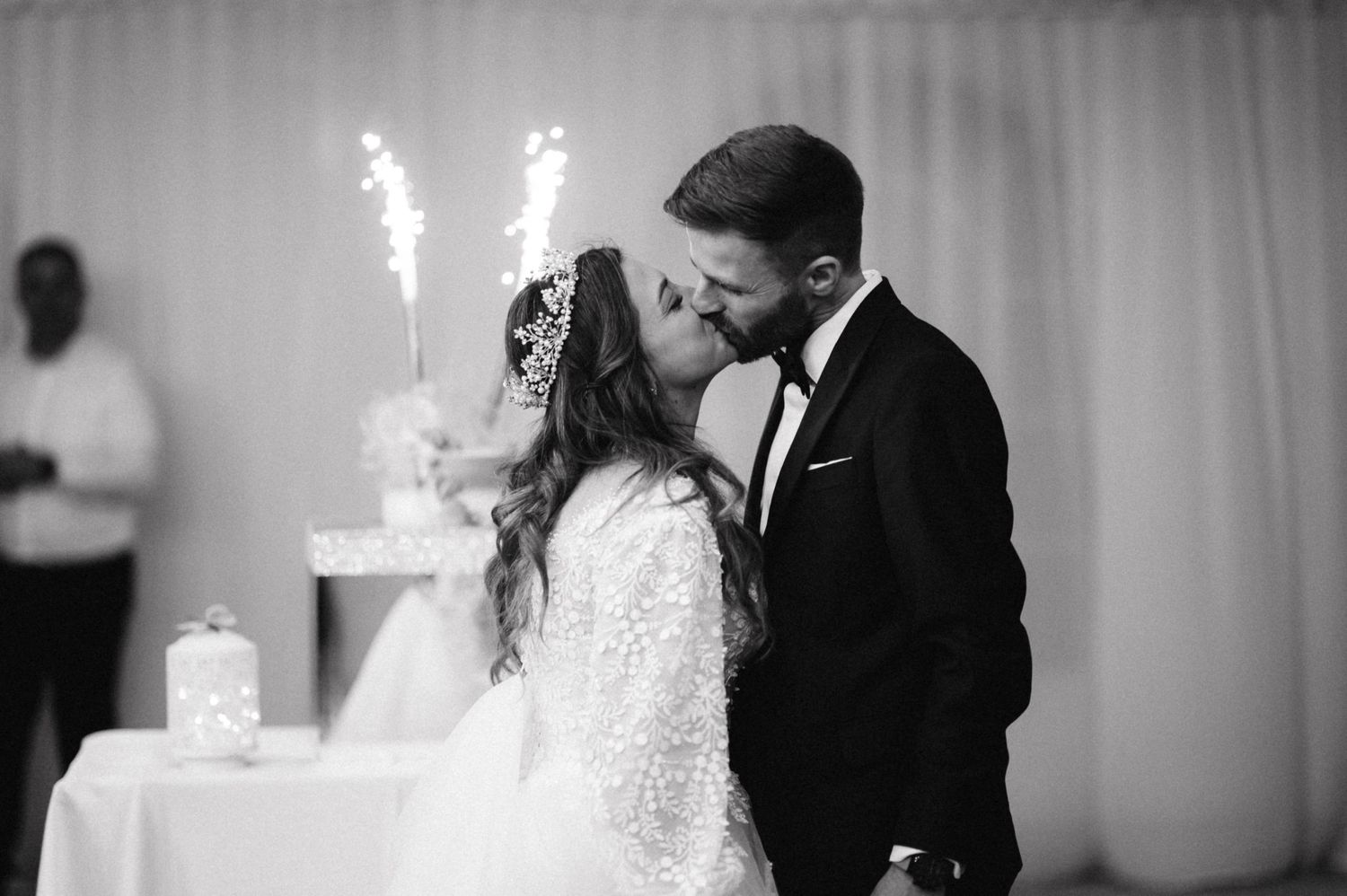 A romantic black and white wedding photo of a couple kissing next to their sparkler-topped cake.