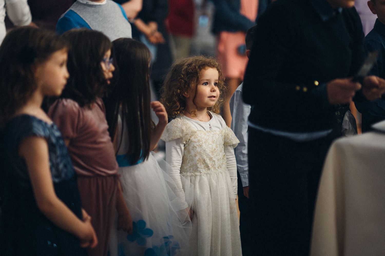Young flower girl in white lace dress stands amid wedding guests.