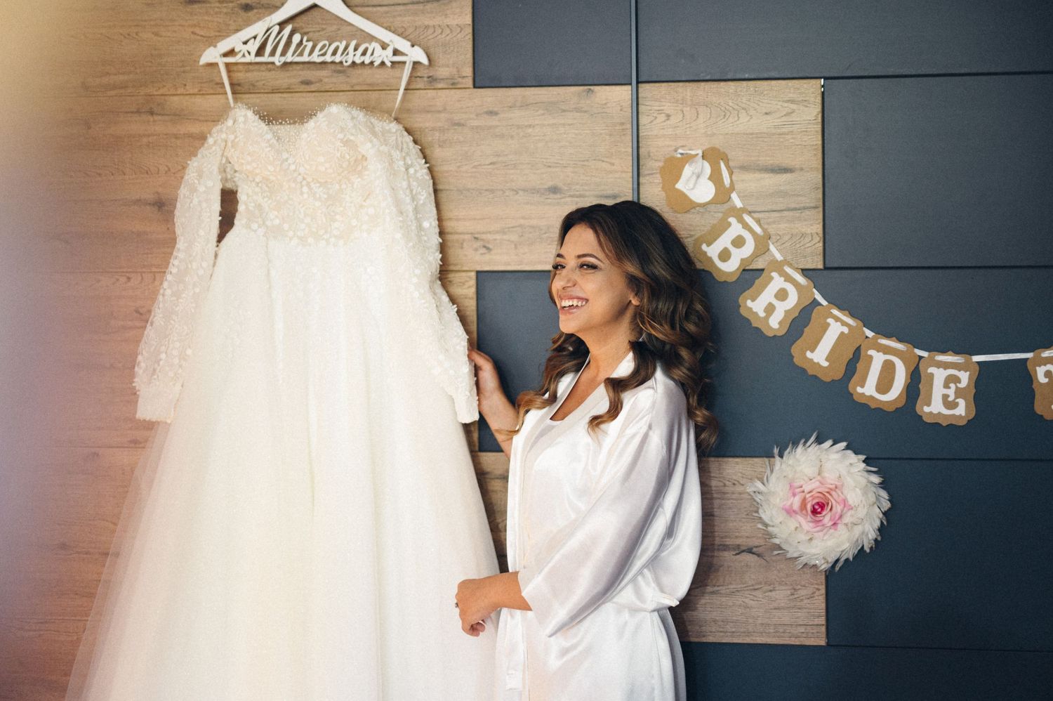 Wedding dress hanging while person in white robe admires it near bride banner decoration.