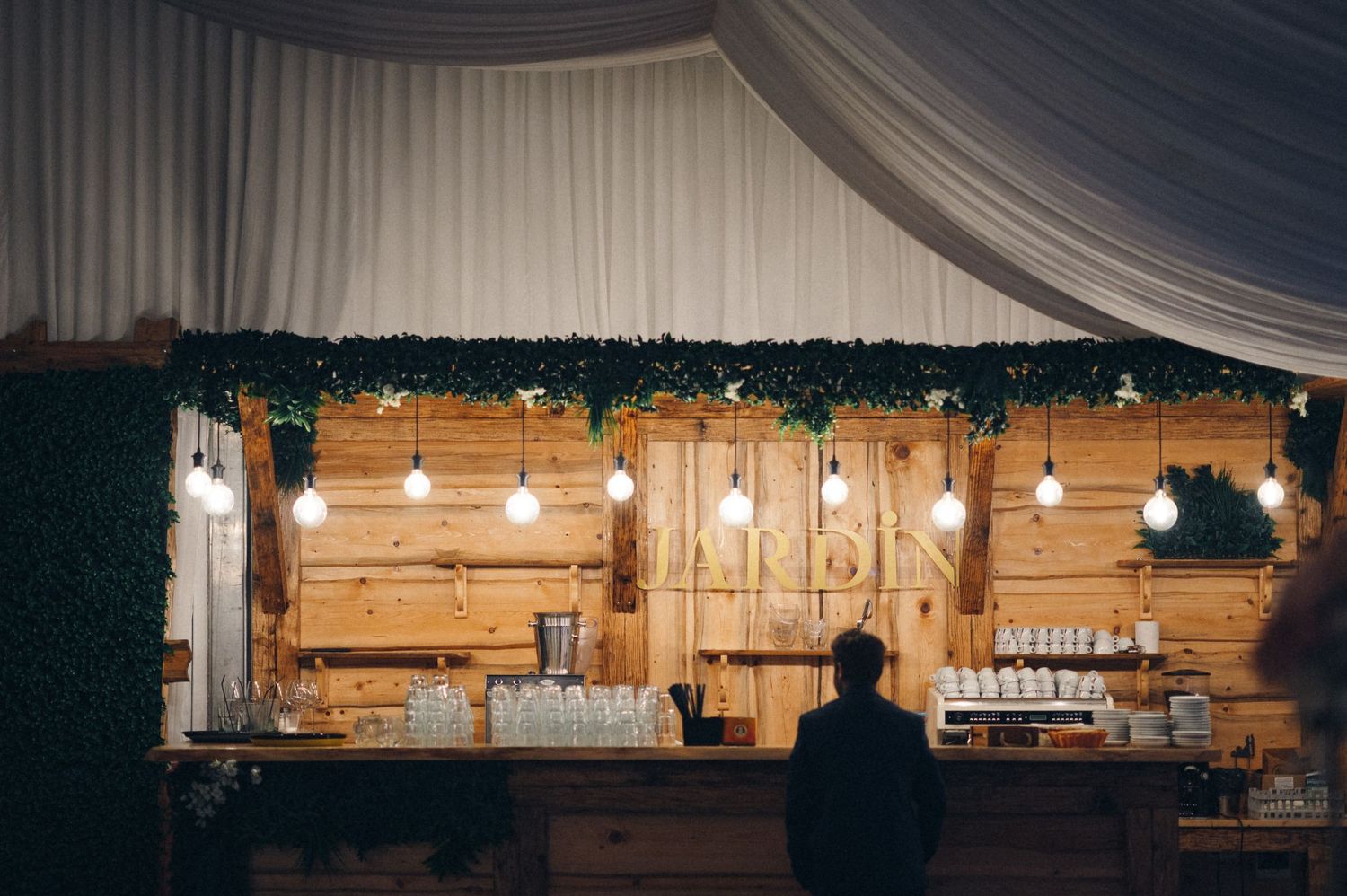 Rustic wooden bar decorated with greenery and string lights at an indoor wedding reception.