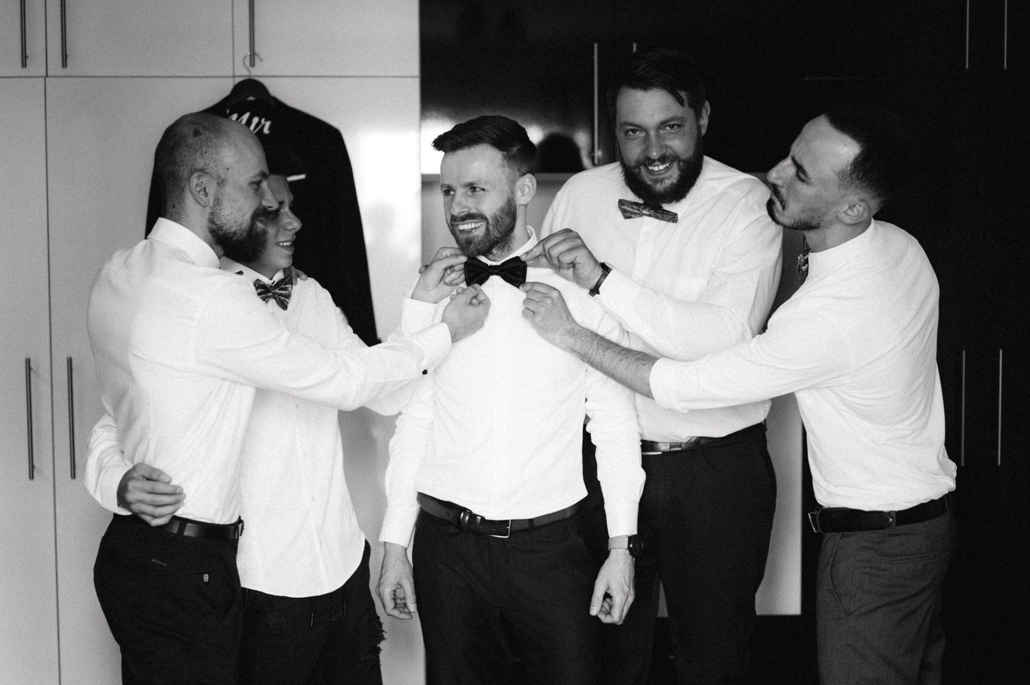 Black and white photo of groomsmen helping adjust formal attire before wedding ceremony.