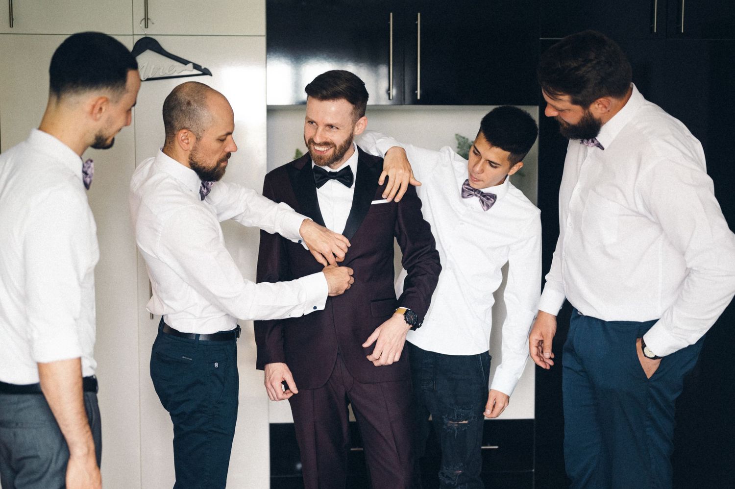 Group gathered around adjusting formal attire in dressing room before ceremony.