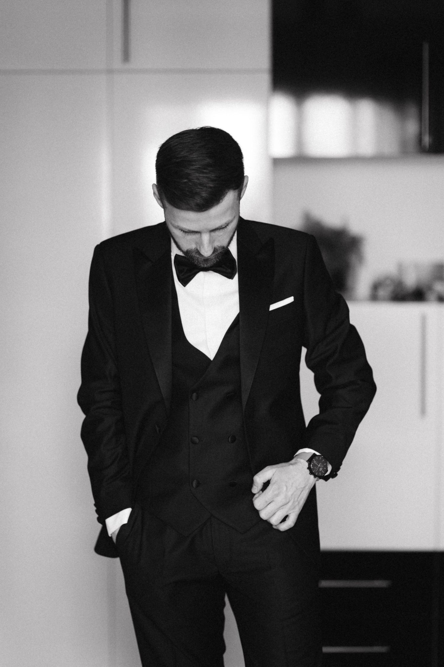 A groom in a classic black tuxedo and bow tie looks down while adjusting his watch in a monochrome photo.