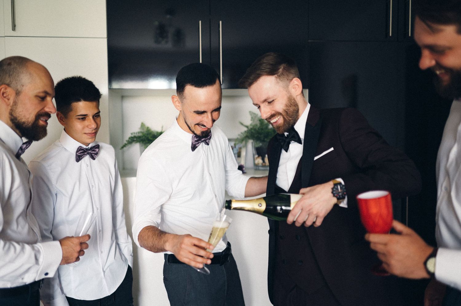 A group of groomsmen share drinks and laughs in a modern kitchen before the wedding ceremony.