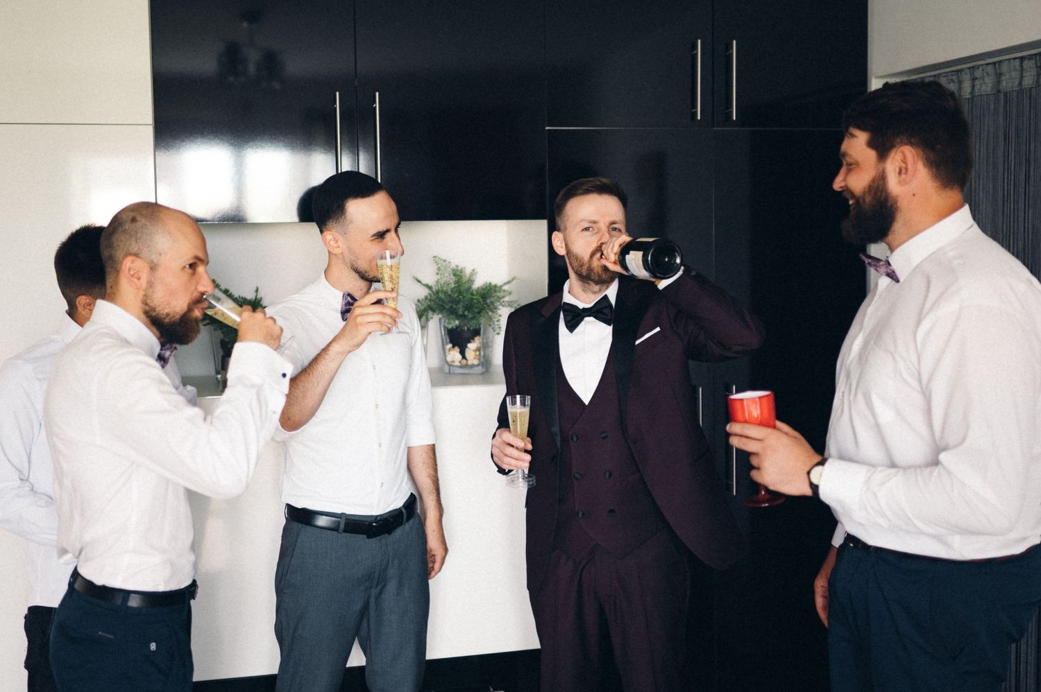 Friends in formal attire enjoying drinks and conversation in a modern kitchen setting.