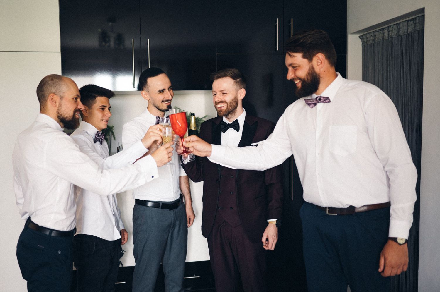 Groomsmen share a celebratory moment with drinks in a modern kitchen before the wedding.