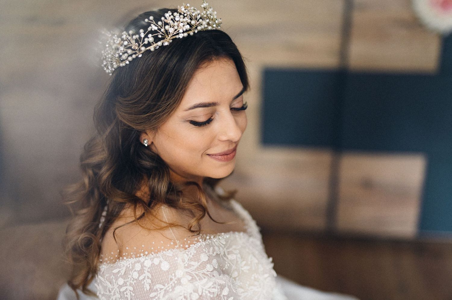 A bride wearing a lace off-shoulder gown and crystal crown smiles gently in a romantic portrait.