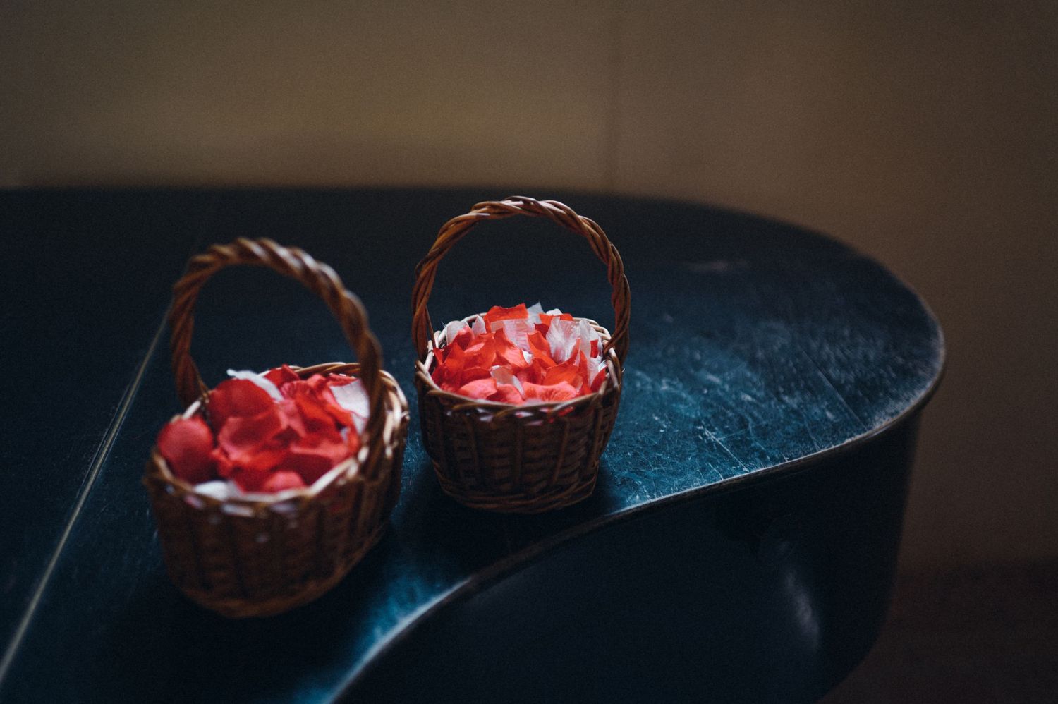Two small woven baskets with red flower petals sit on a dark surface.