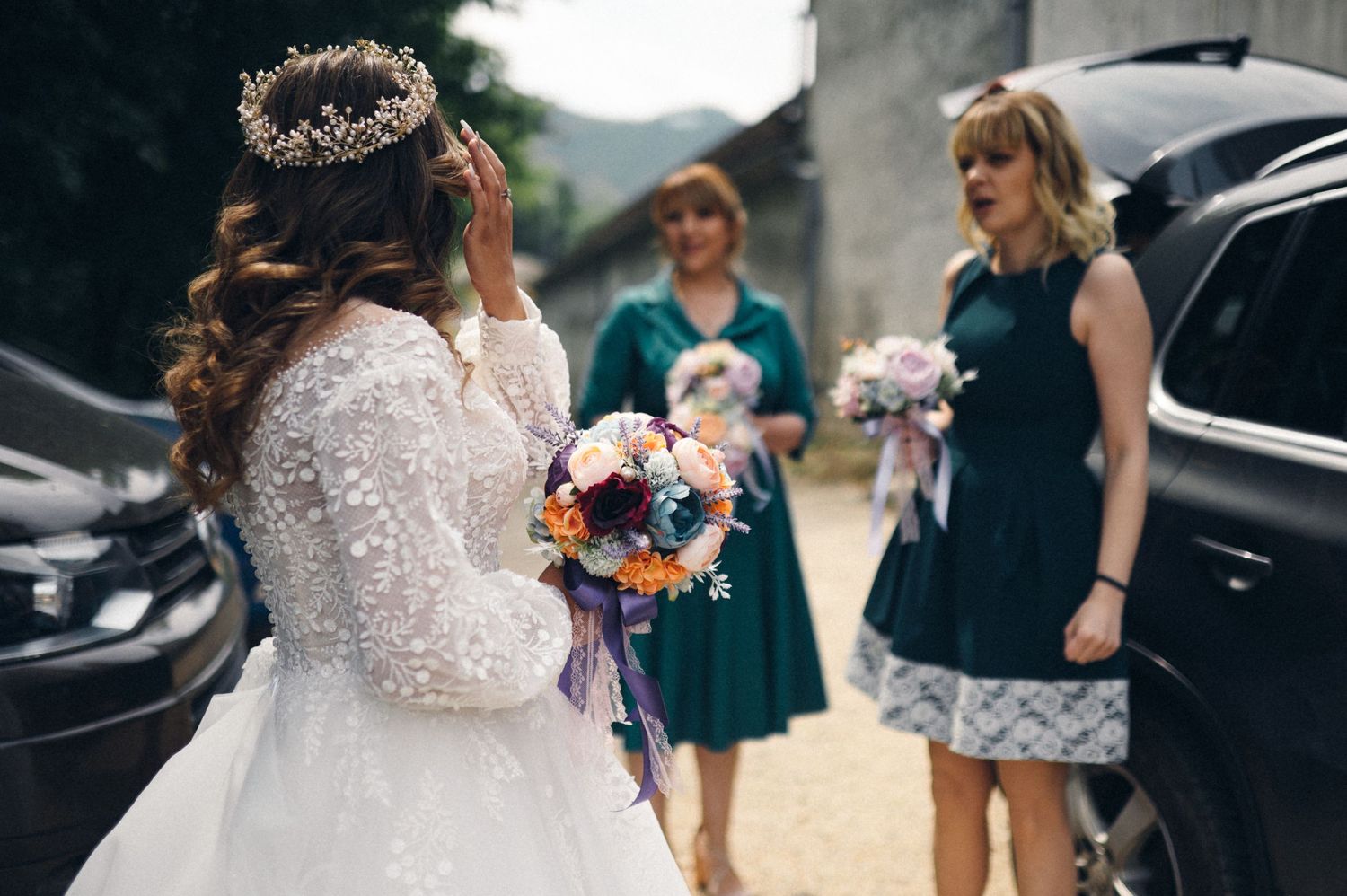 A wedding scene outdoors with attendees in formal wear gathered near parked cars.