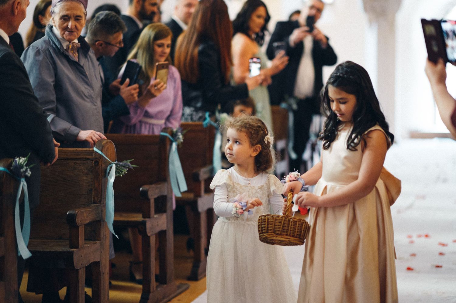 Young flower girls in white dresses walk down the church aisle with baskets.