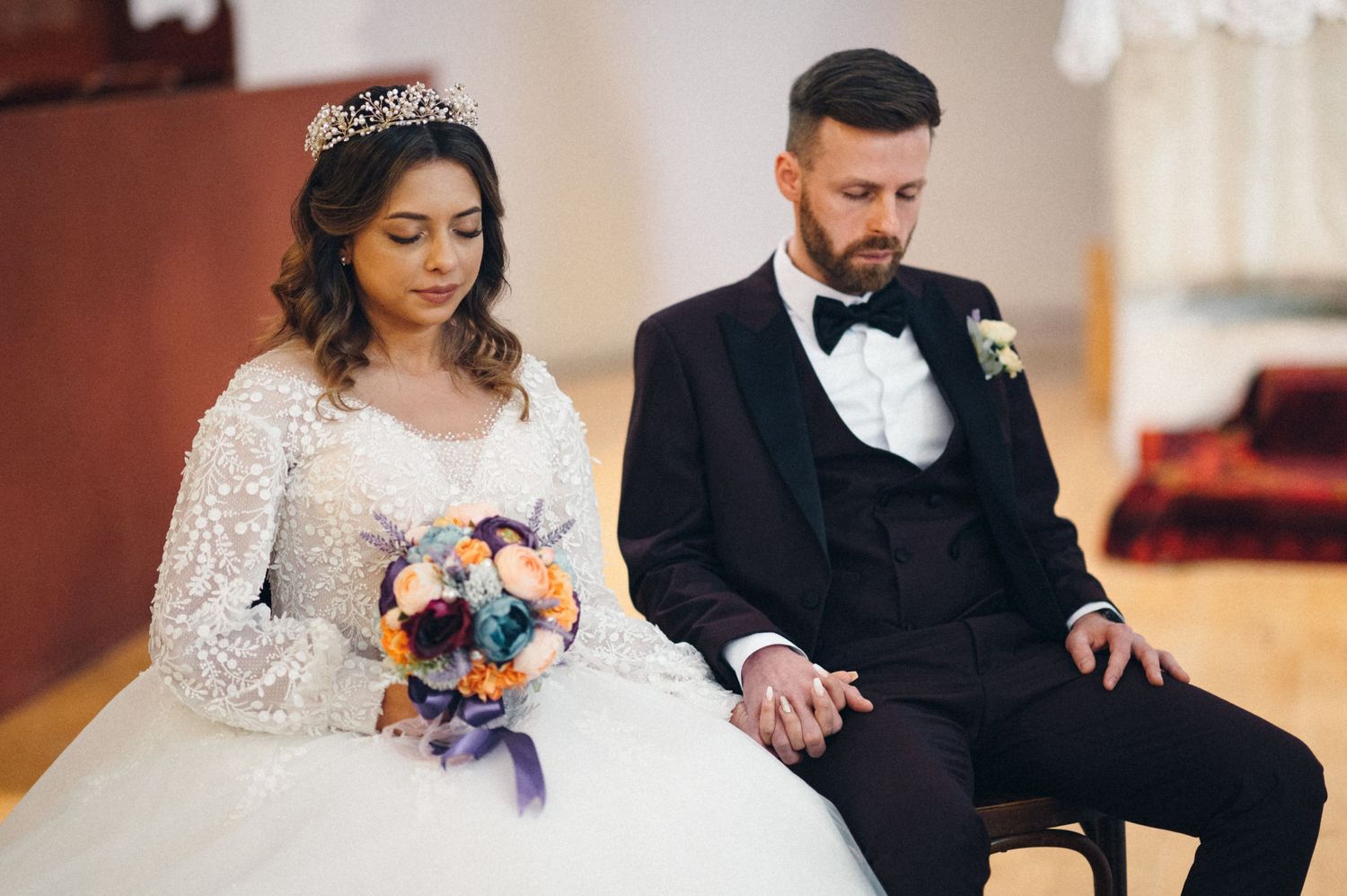 A wedding couple sits together during their ceremony with the bride holding a colorful bouquet.
