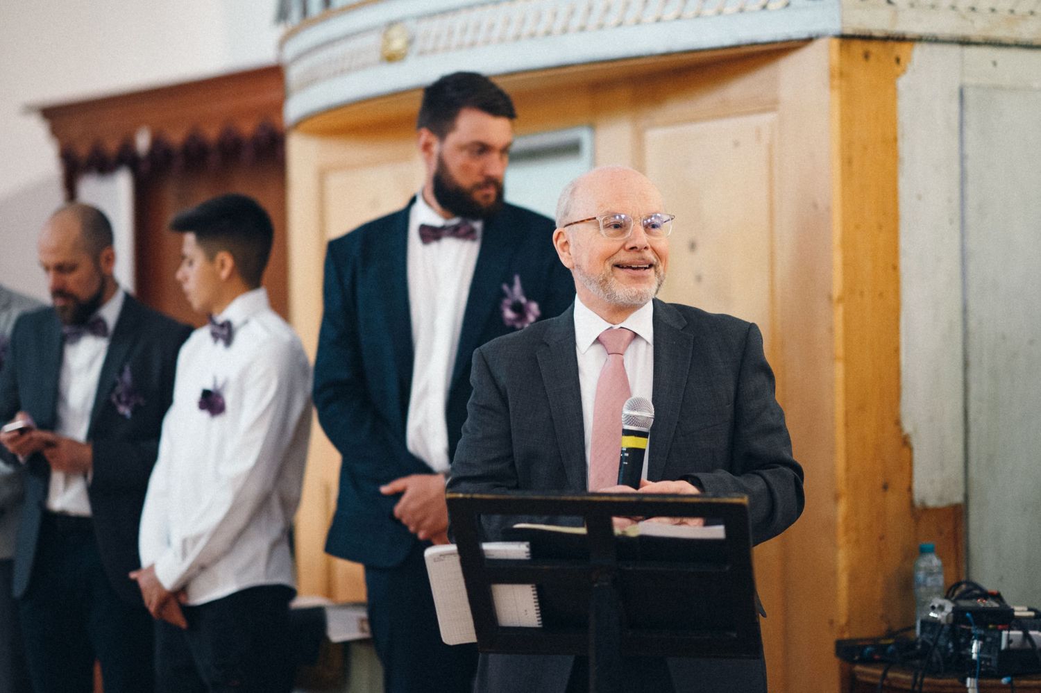 A speaker in a suit and pink tie addresses the audience at a formal event.