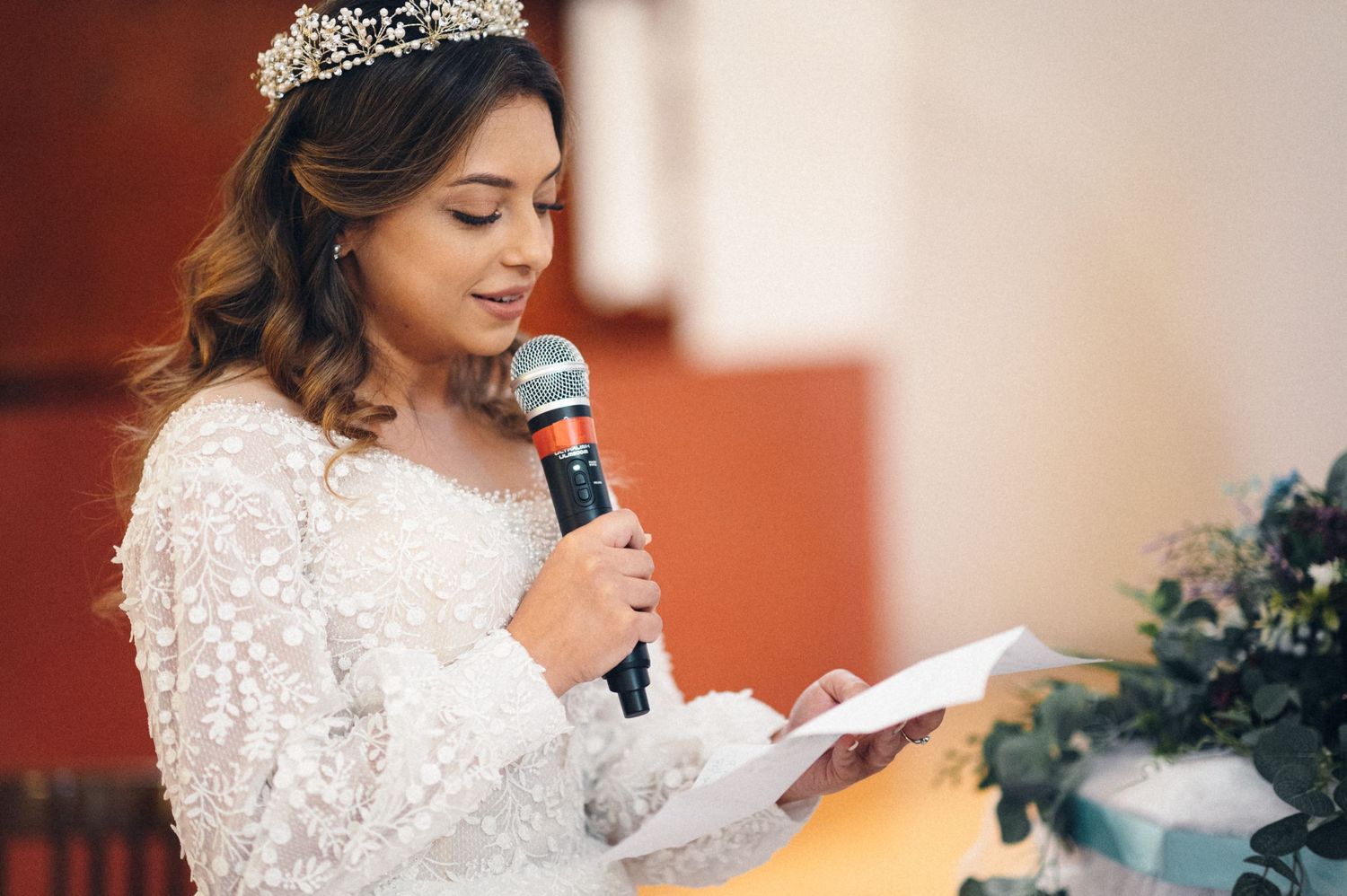 Bride in a lace dress and tiara reads from paper while speaking into a microphone.