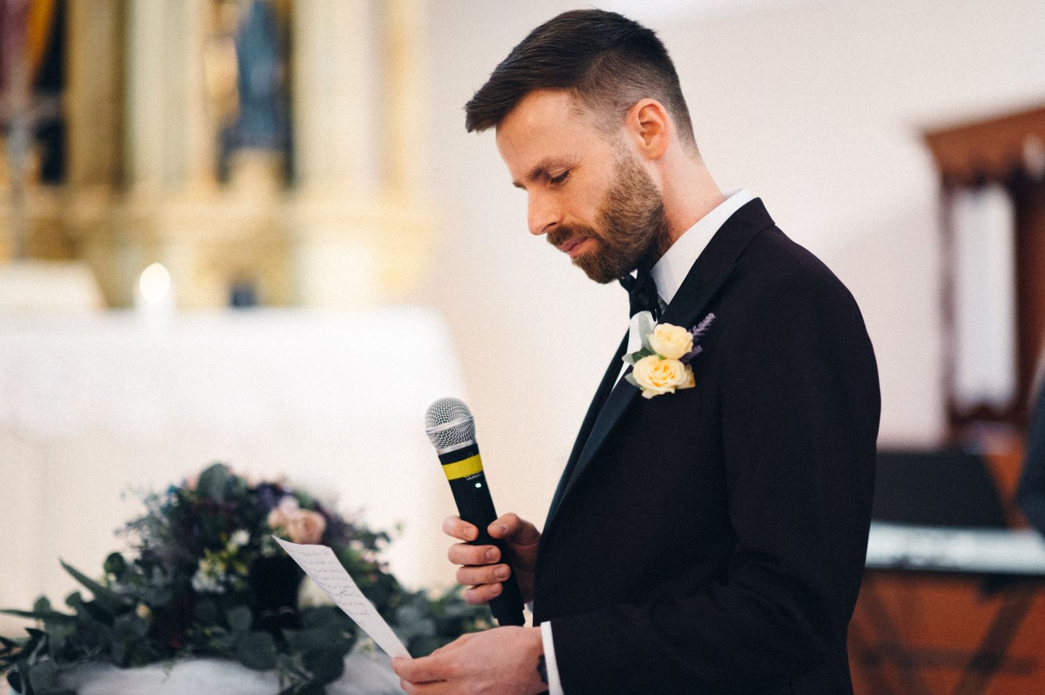 Groom in black tuxedo speaks into microphone during wedding ceremony.