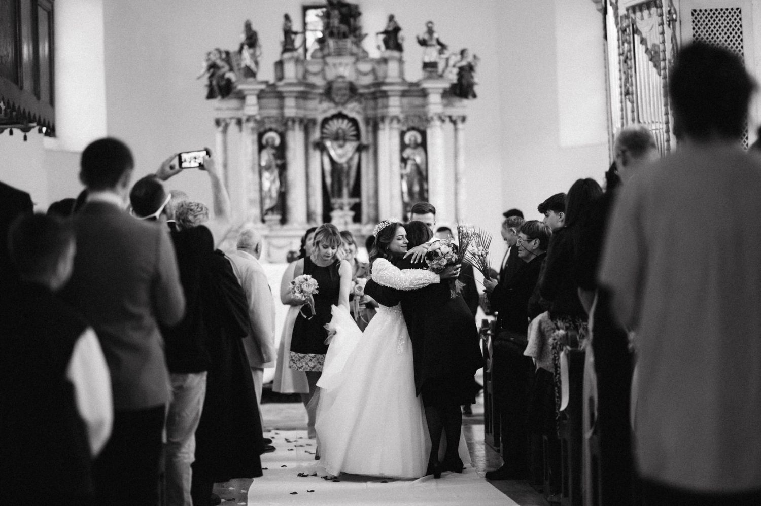 A black and white photo of newlyweds embracing while guests look on in a church aisle.
