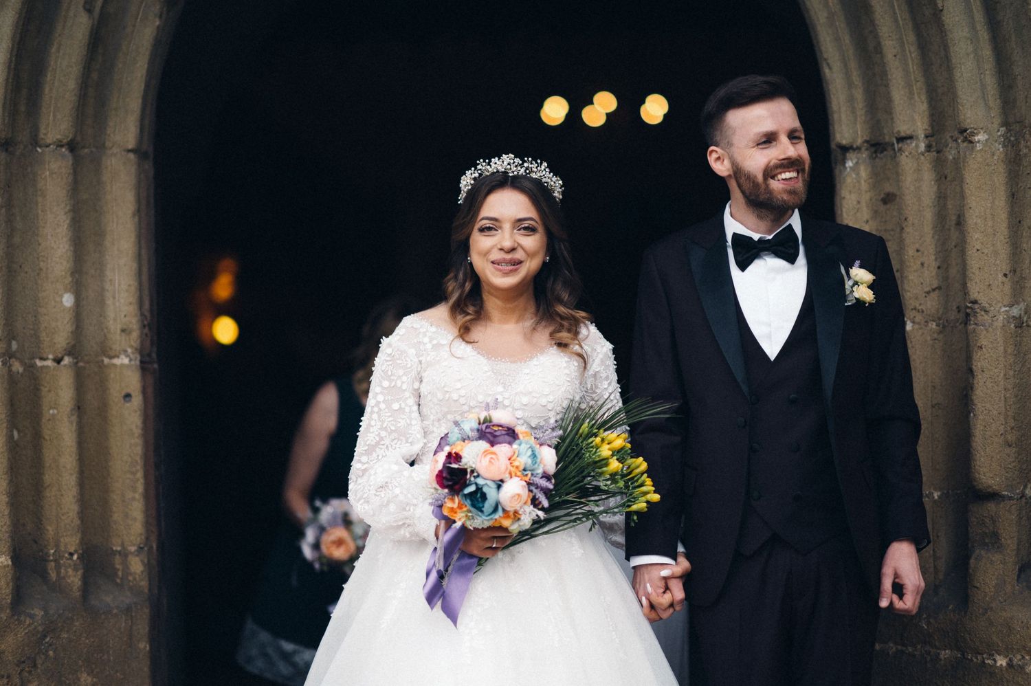 Wedding couple standing under stone church archway holding colorful bouquet.