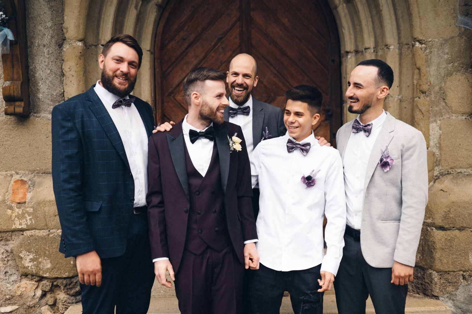 Groomsmen in formal attire posing together outside stone church doorway.