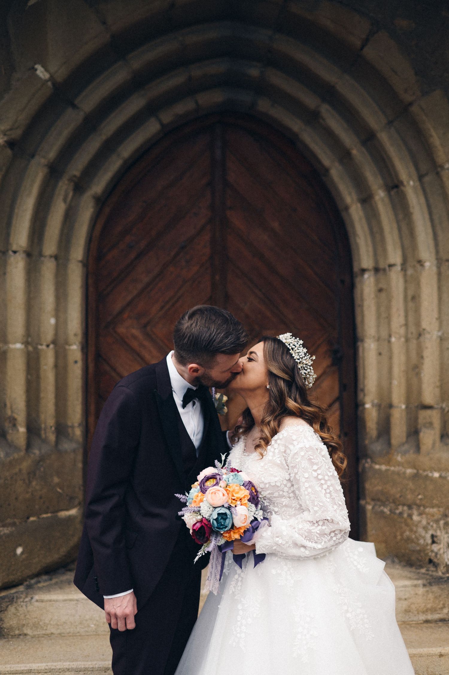 Romantic moment captured between newlyweds with colorful bouquet under stone archway.