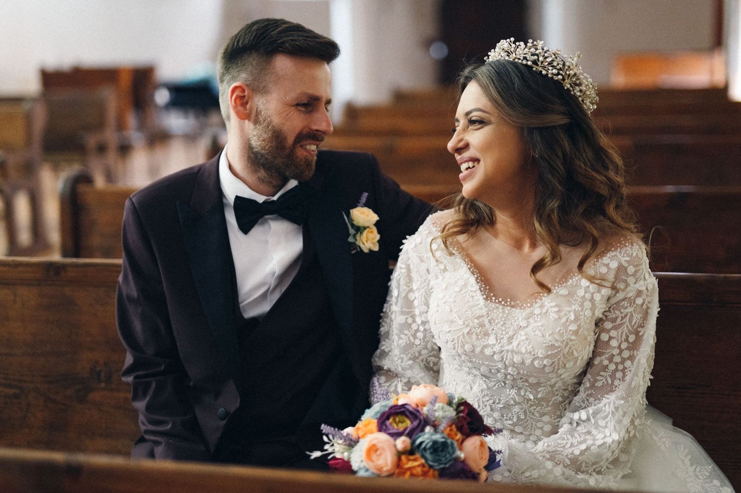 Joyful wedding couple sharing laughter in church pews with floral bouquet.
