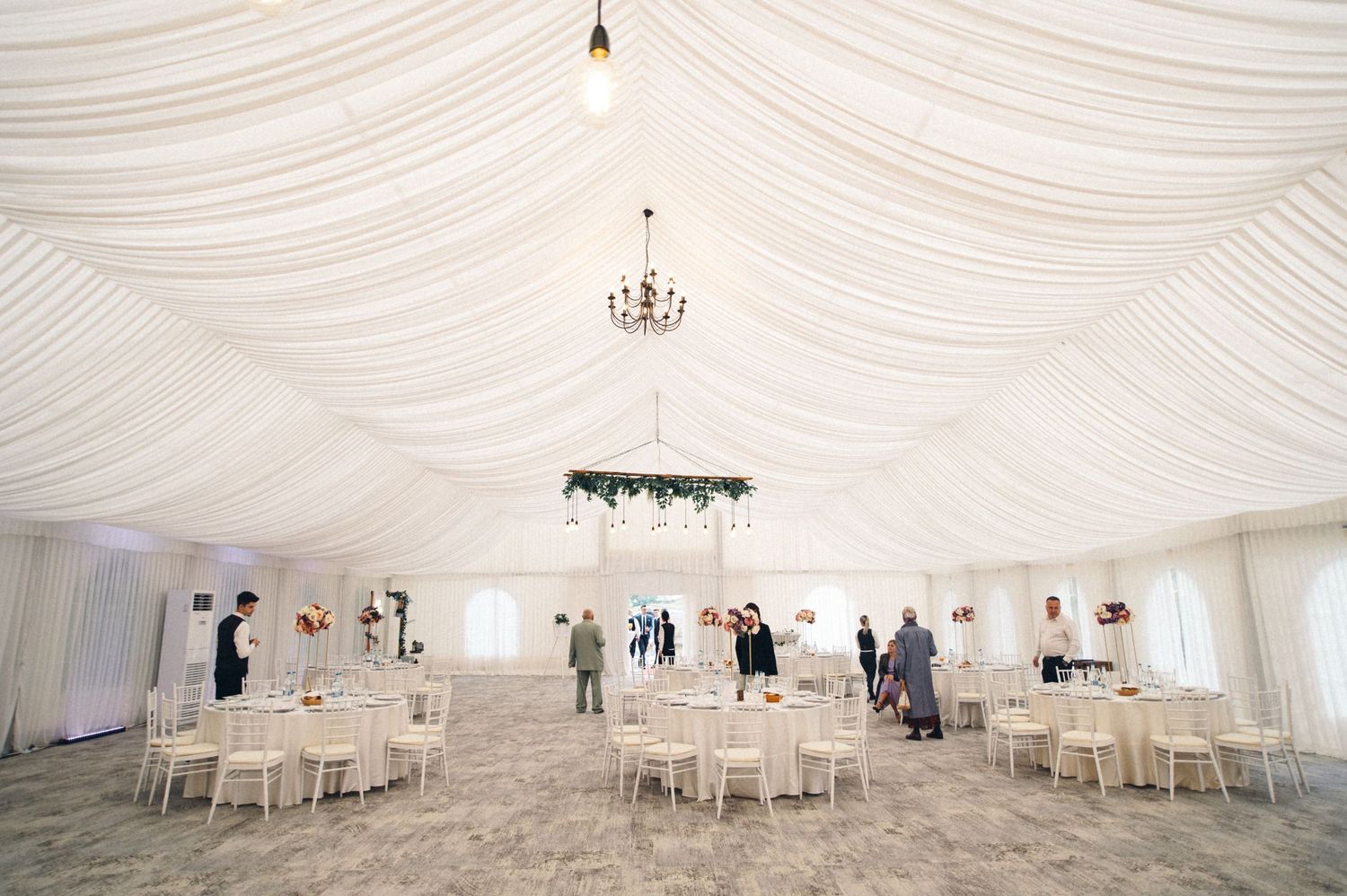 Spacious white wedding tent interior with draped ceiling, chandeliers and round table settings.