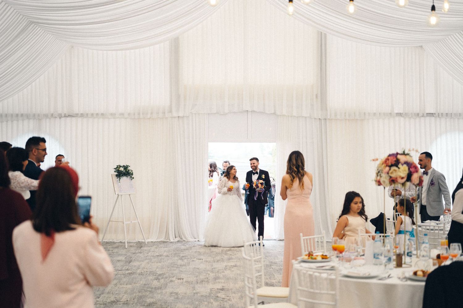 Wedding reception in progress inside an elegant white tent with string lights and dining tables.