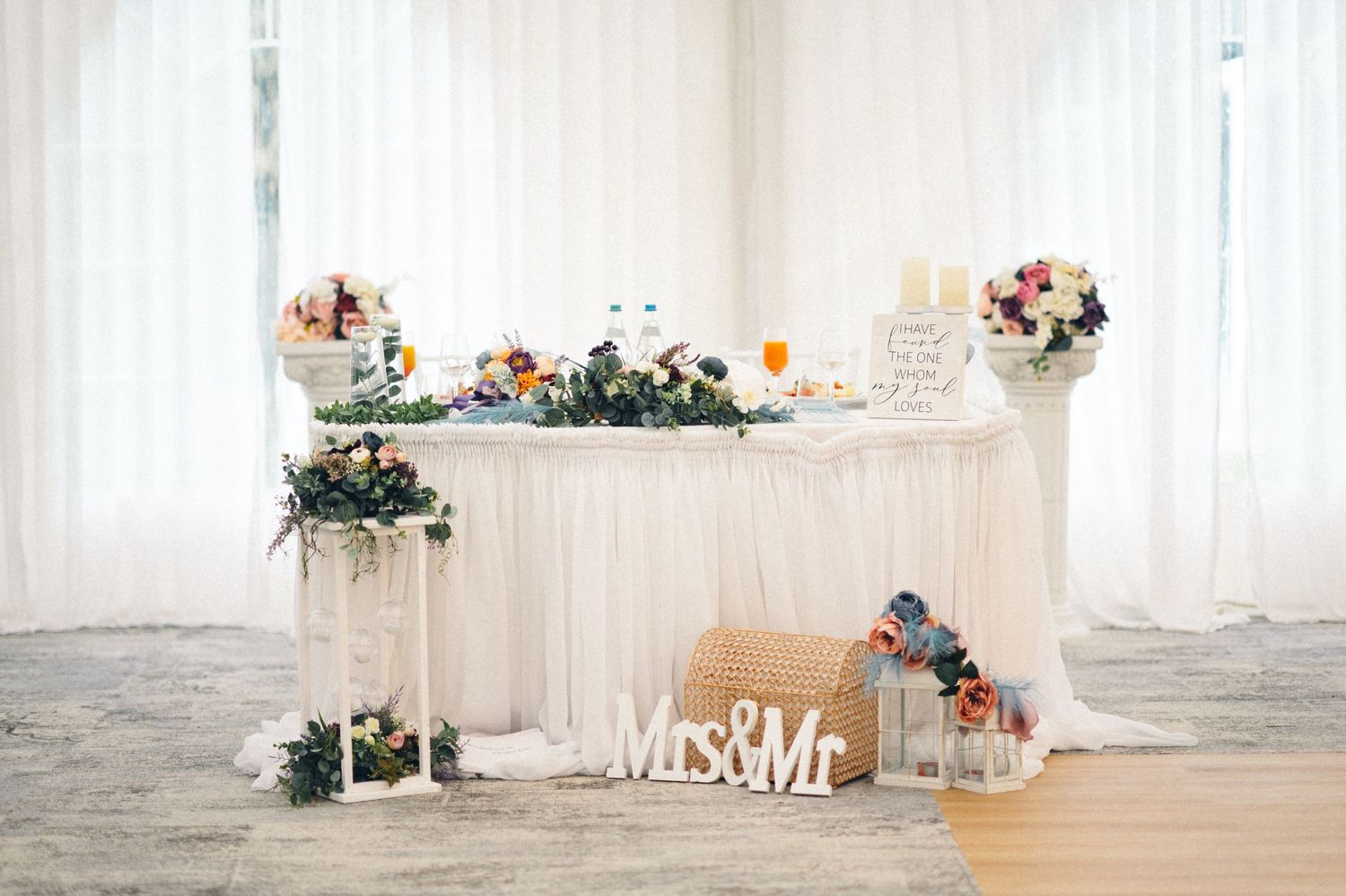 White wedding dessert table decorated with flowers, Mr & Mrs sign, and elegant decor pieces.