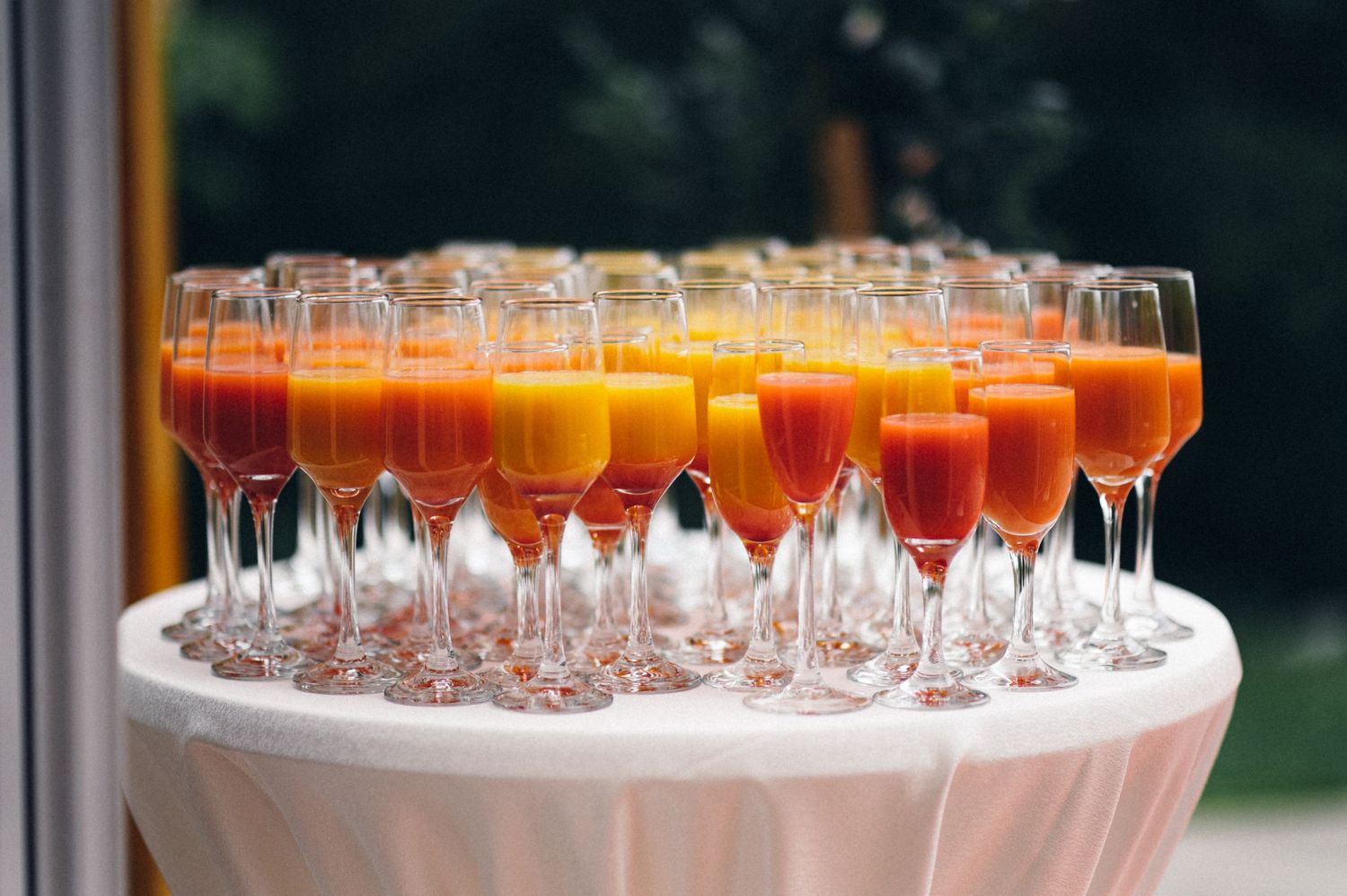 Champagne flutes filled with colorful cocktails arranged on a round white table at an outdoor event.