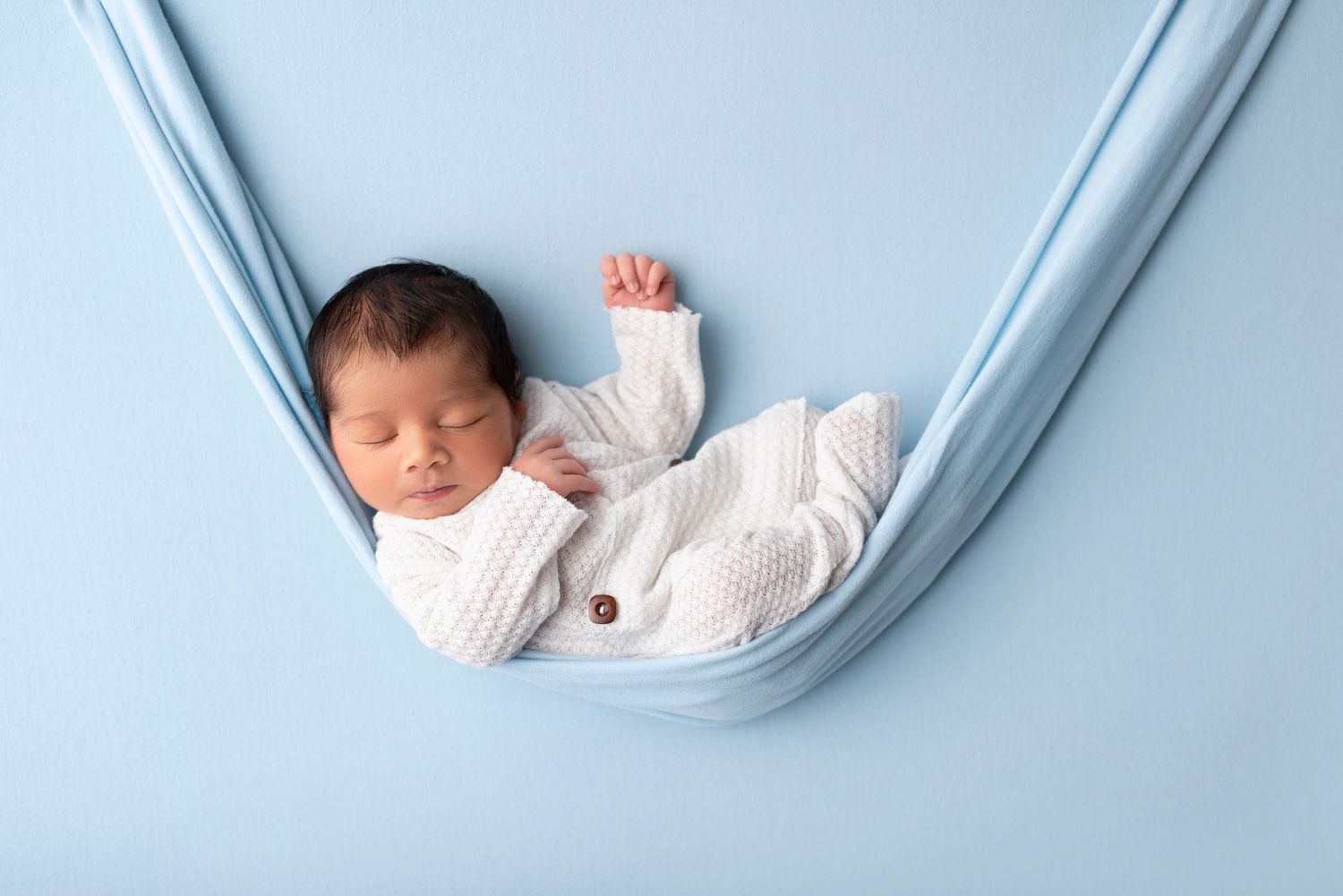 Newborn baby sleeping peacefully in white outfit on blue fabric hammock.