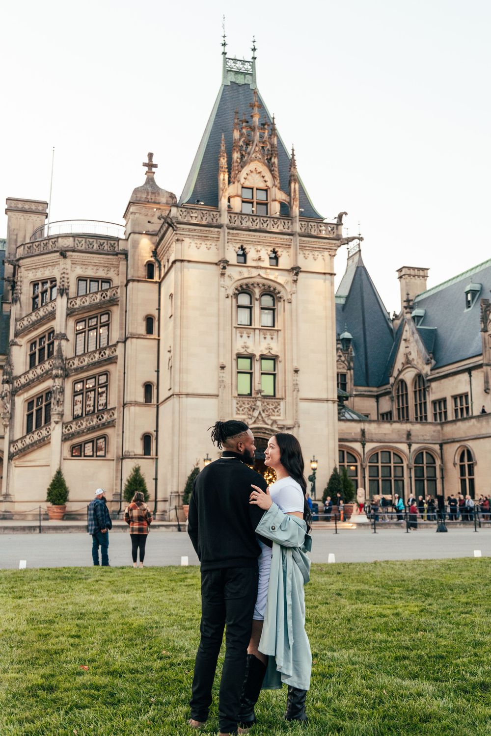 A couple stands on grass in front of the majestic Biltmore Estate in Asheville.