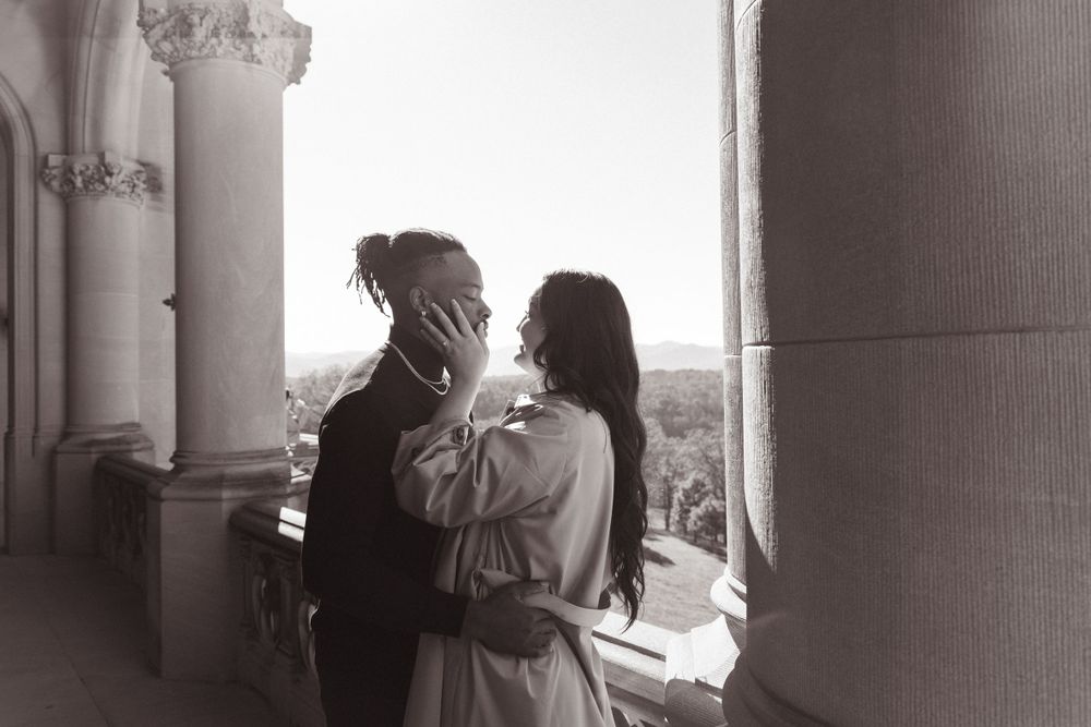 Black and white photo of a romantic embrace on a historic balcony with scenic background.