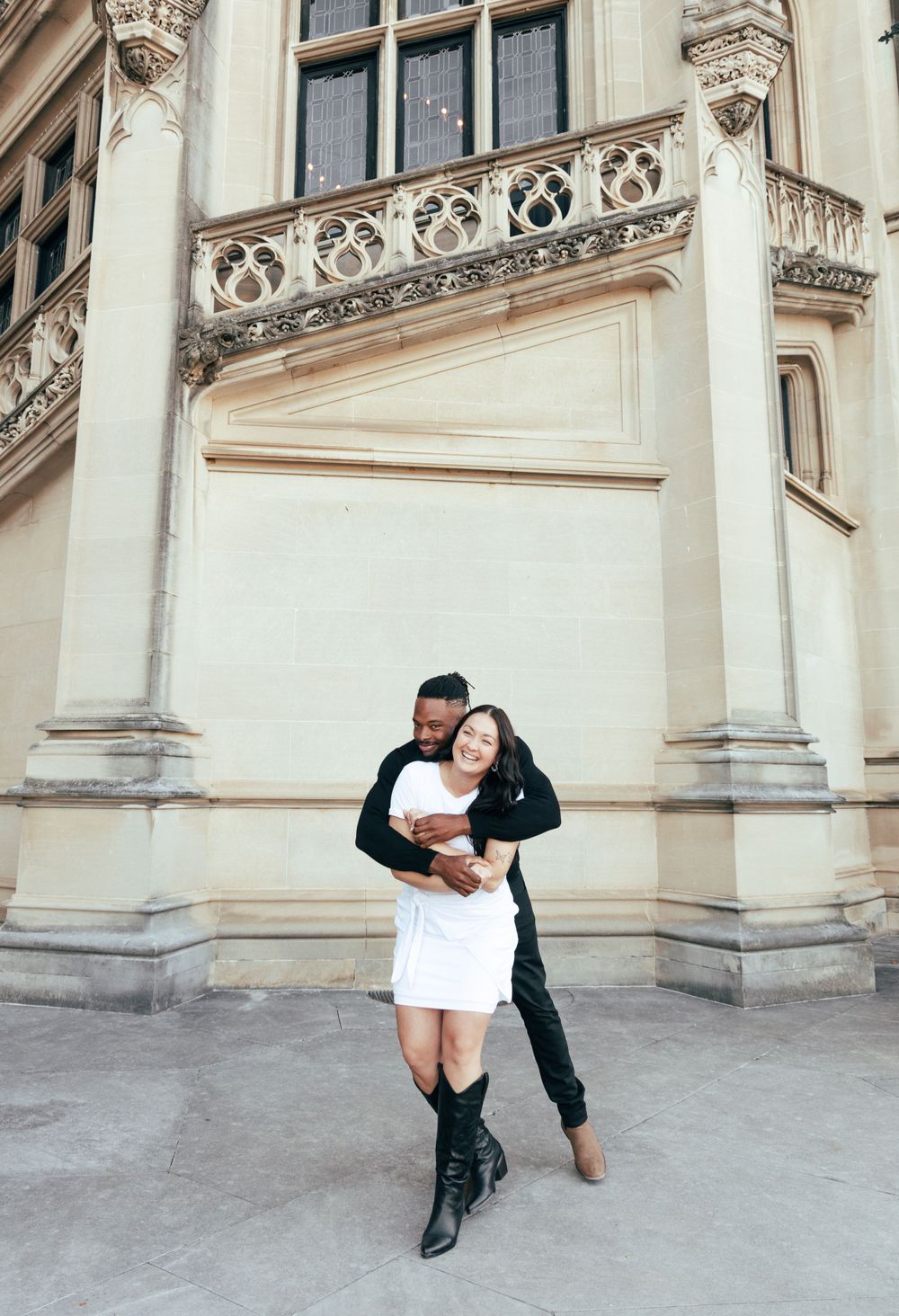 Playful couple embracing in front of classical architecture with ornate stone details.