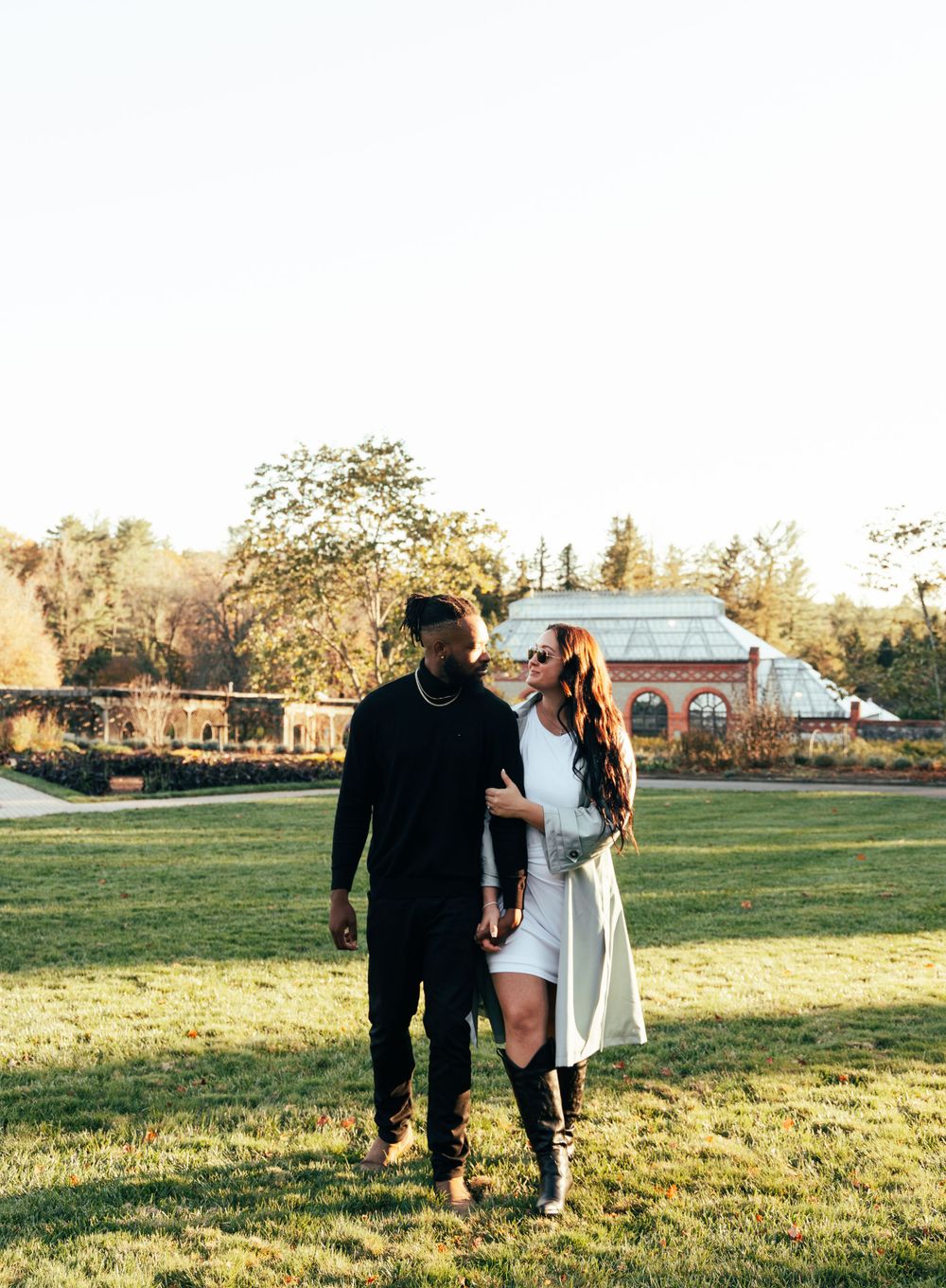 Couple walking together on grass during golden hour with historic buildings in background.