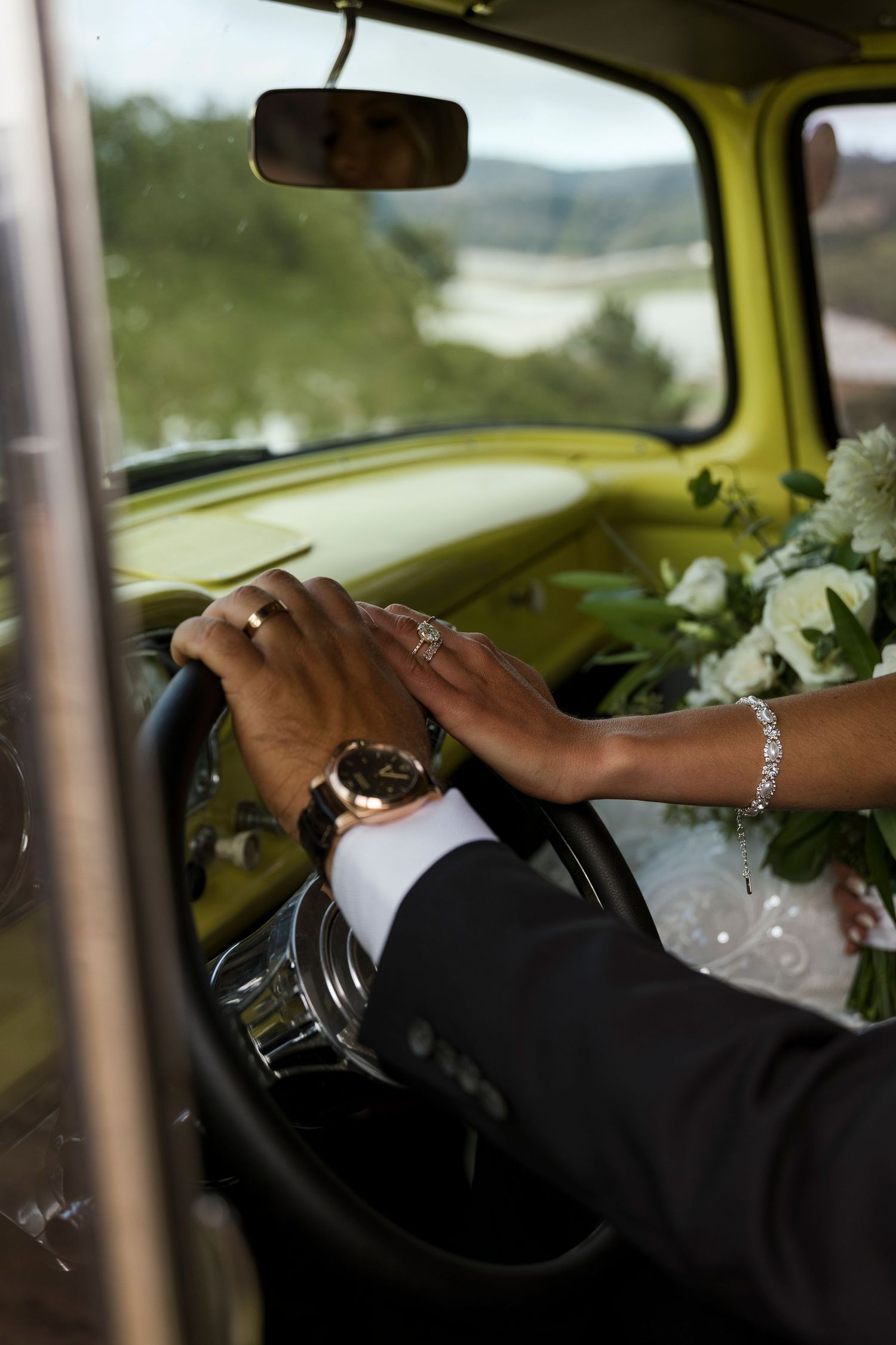 Close-up of hands on steering wheel in classic car with wedding flowers visible.