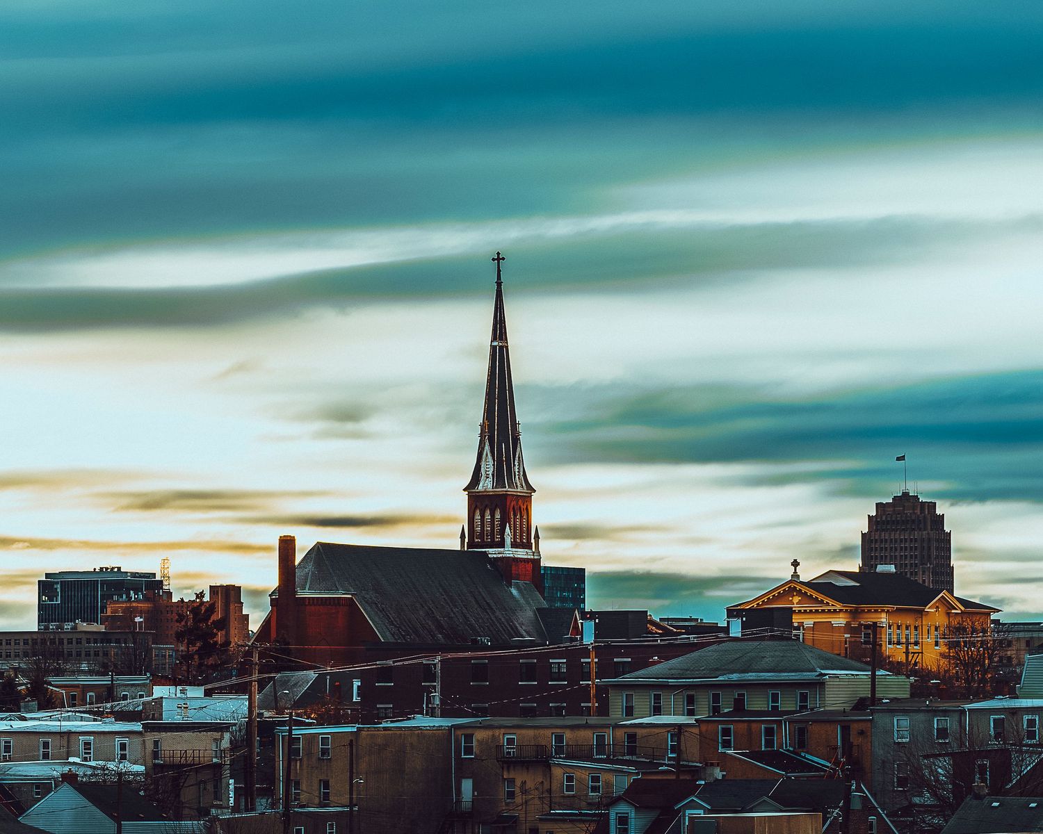 Historic city skyline with church spire and buildings against a dramatic turquoise sky at dusk.