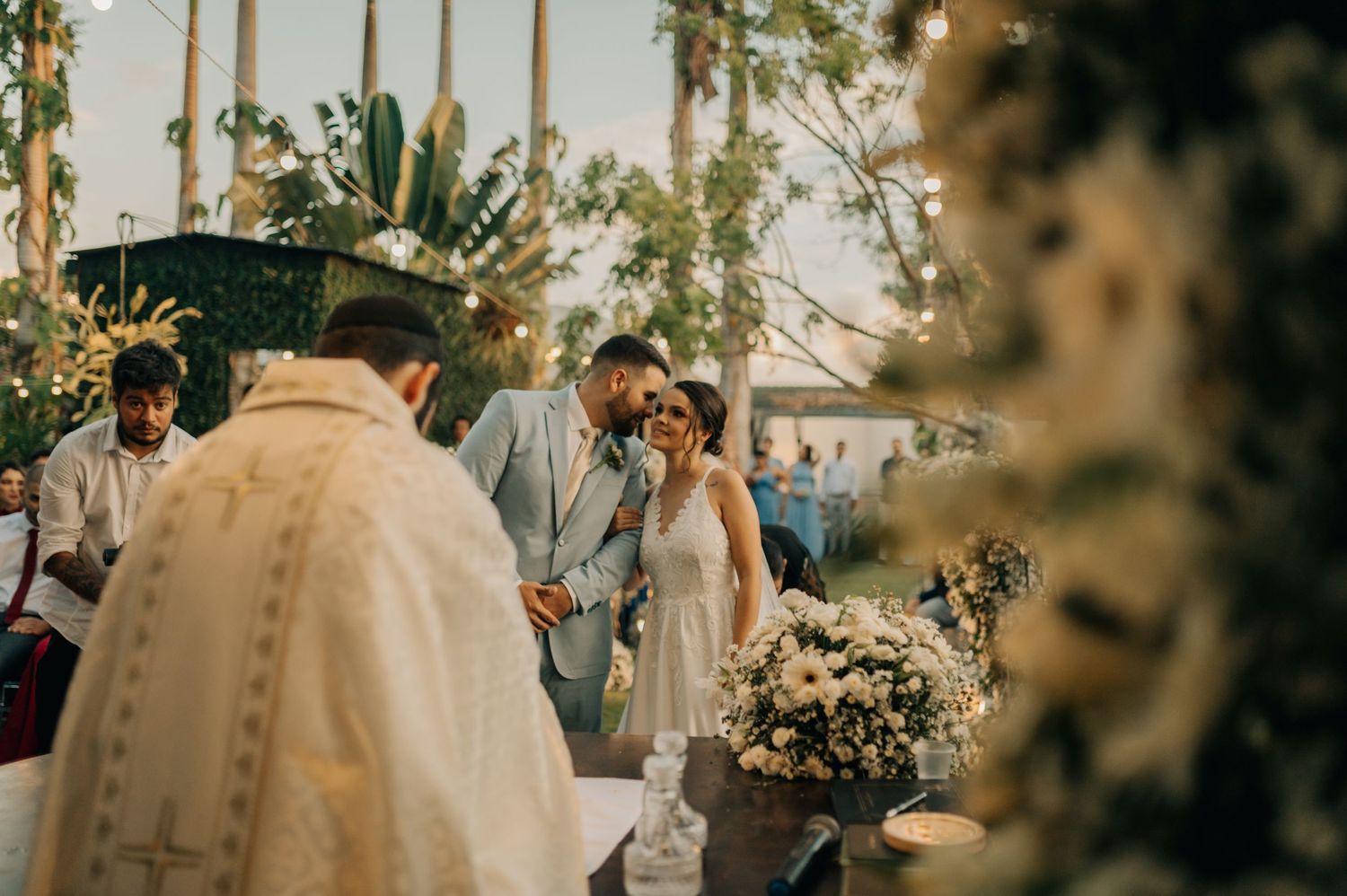 An intimate outdoor wedding ceremony with floral decorations and palm trees in the background.