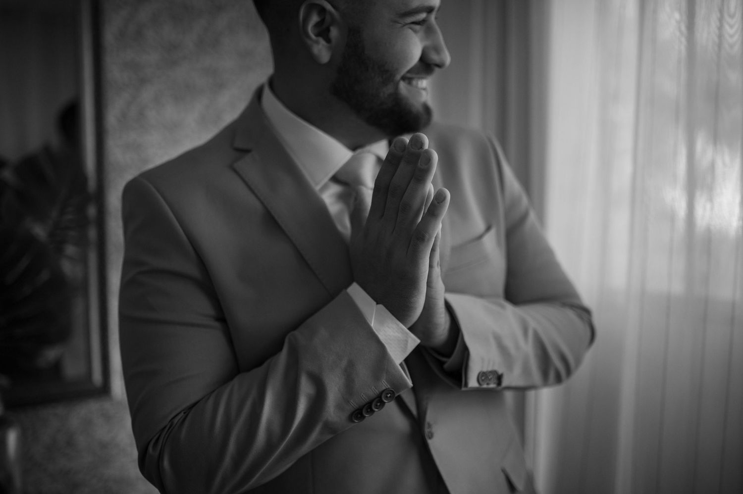 A groom in a gray suit stands in contemplative prayer by a window.