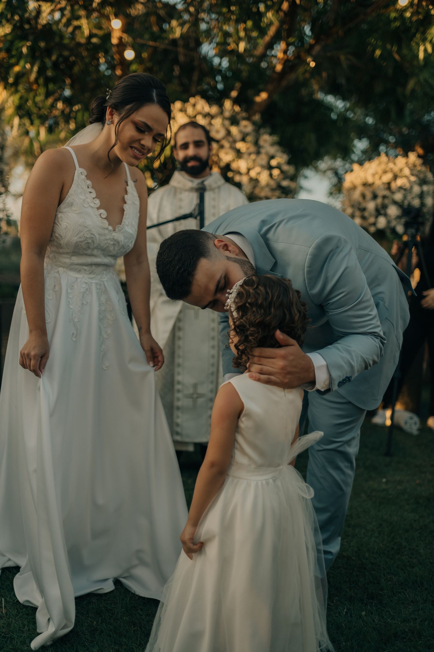 Tender moment between wedding couple and flower girl during outdoor ceremony.