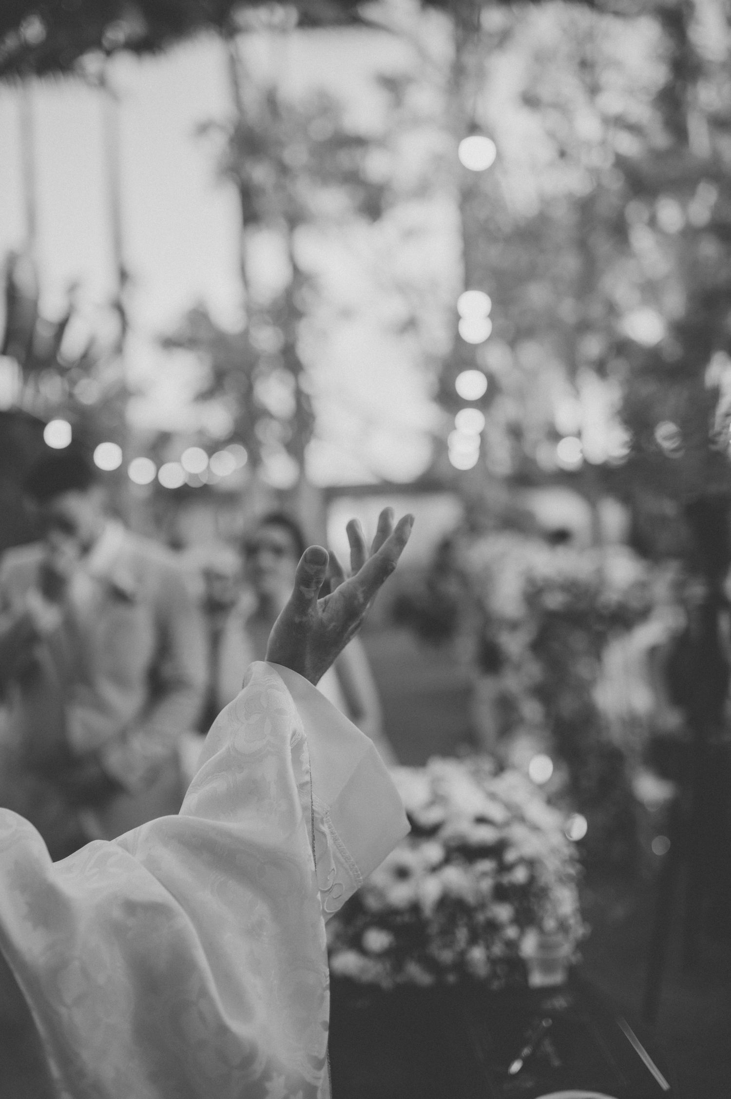 A raised hand in white sleeve against bokeh lights during an outdoor ceremony.