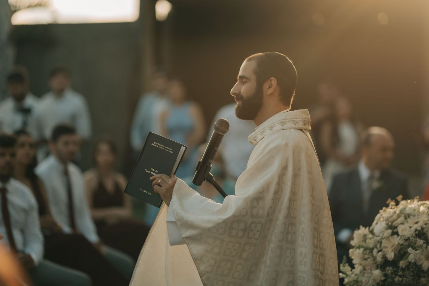 A priest in white robes holds a microphone and book during an outdoor wedding ceremony.