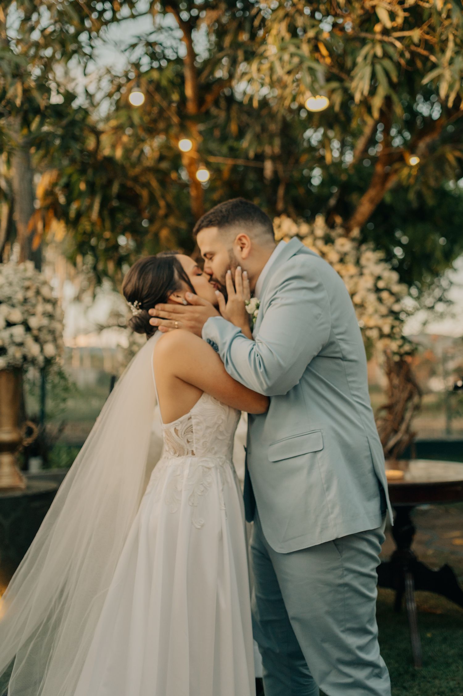 Romantic first kiss as newlyweds under trees with string lights at outdoor wedding.