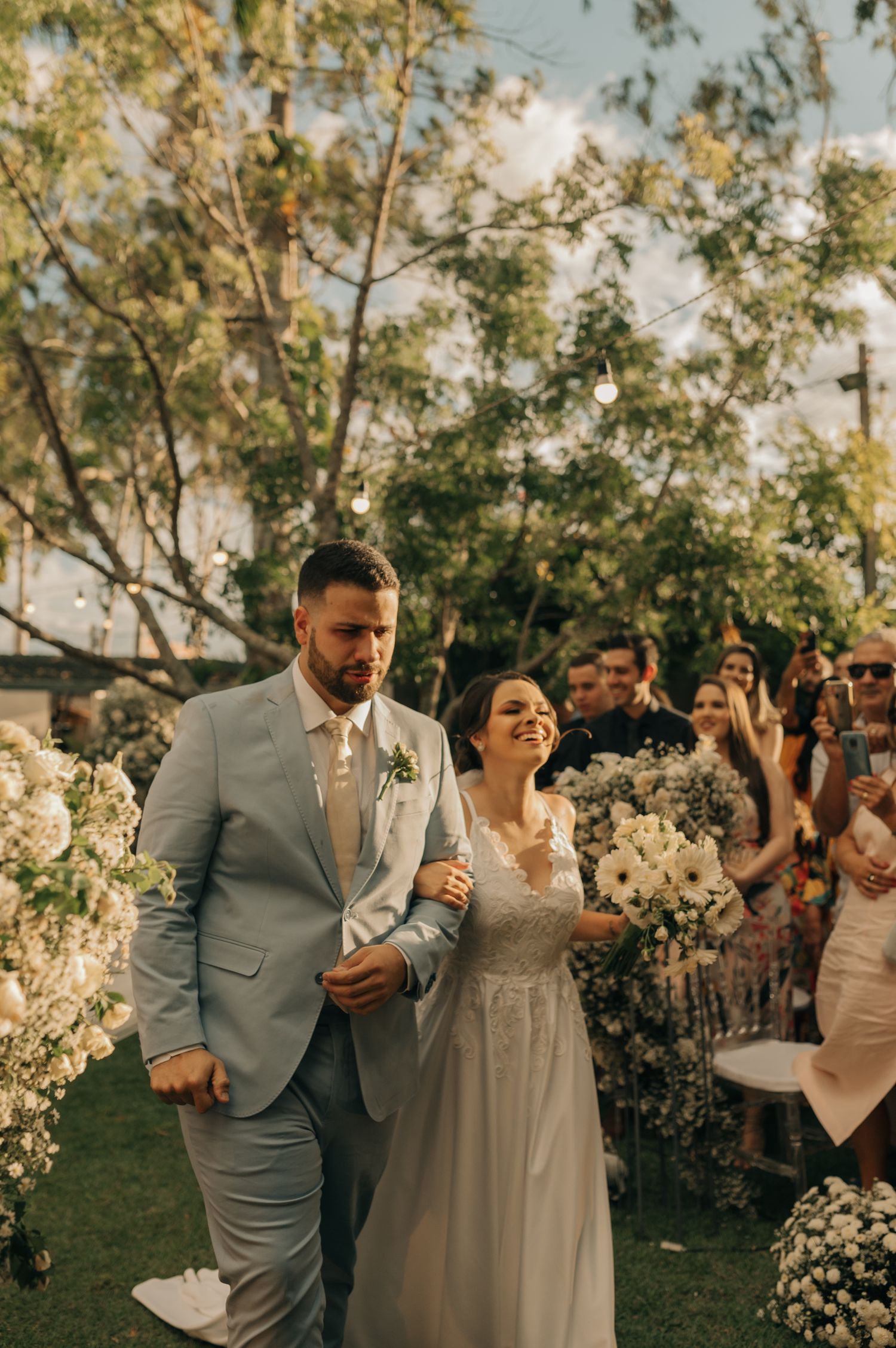 Newlyweds walk down the aisle as wedding guests celebrate in an outdoor garden ceremony.