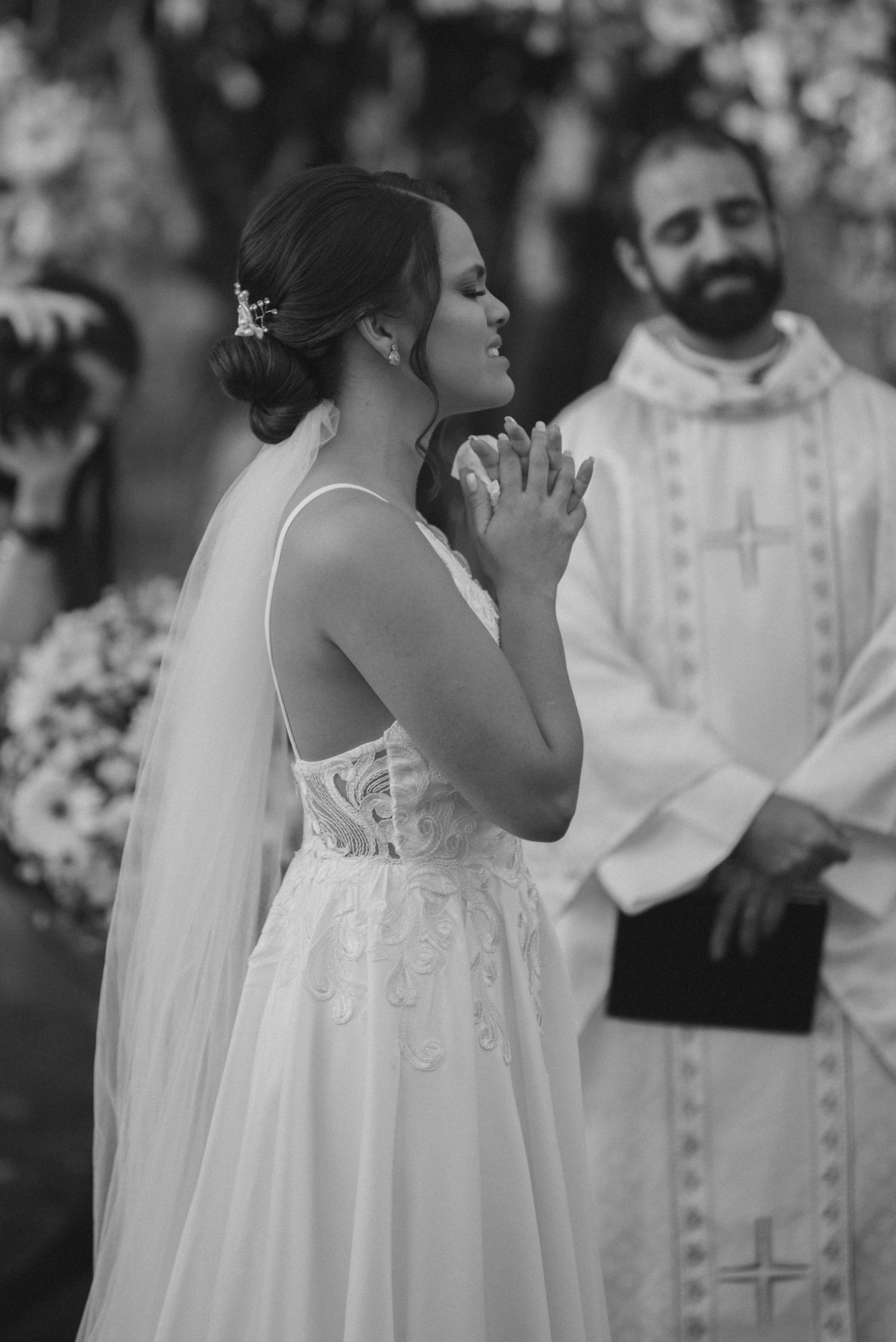 A bride in a white lace dress clasps her hands in prayer during the wedding ceremony.