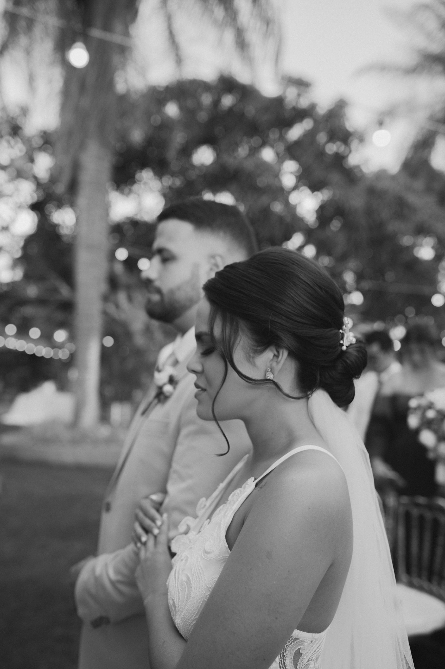 Black and white outdoor wedding portrait with palm trees and string lights in background.
