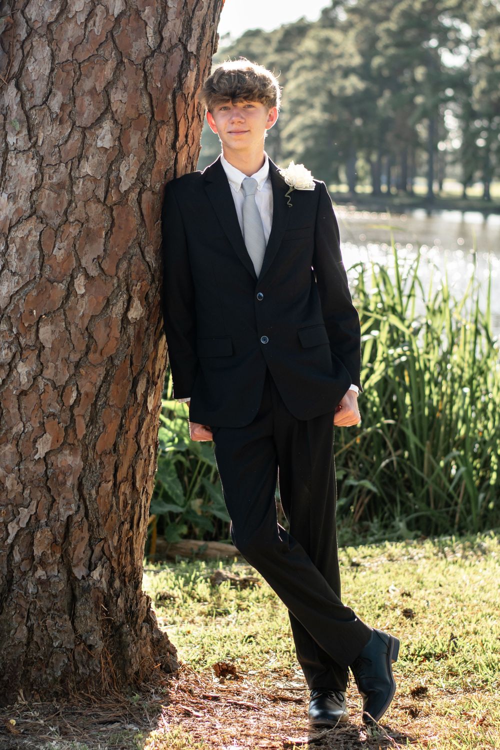 A formal portrait of someone in a black suit leaning against a large pine tree.