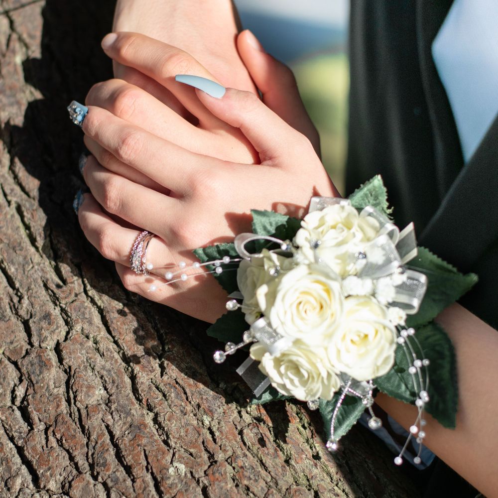 A white rose corsage and engagement ring shown on hands against tree bark.