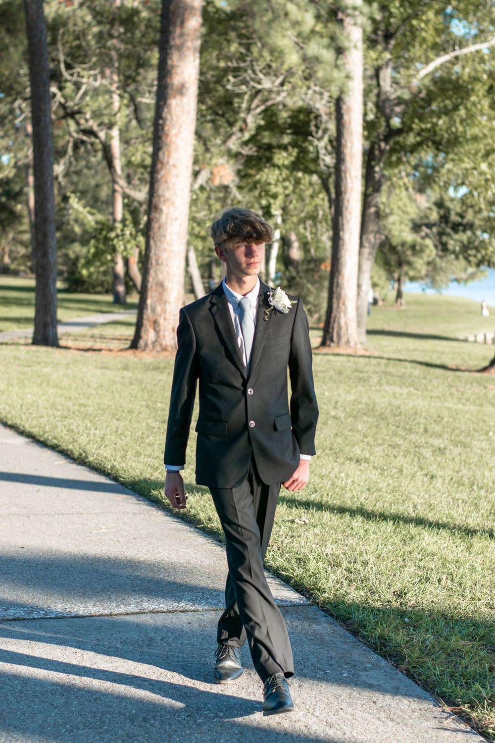 Person in formal black suit and tie walks along a tree-lined path in a park.