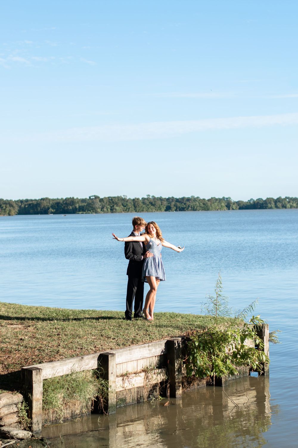 A couple stands with arms outstretched on a dock by a lake.