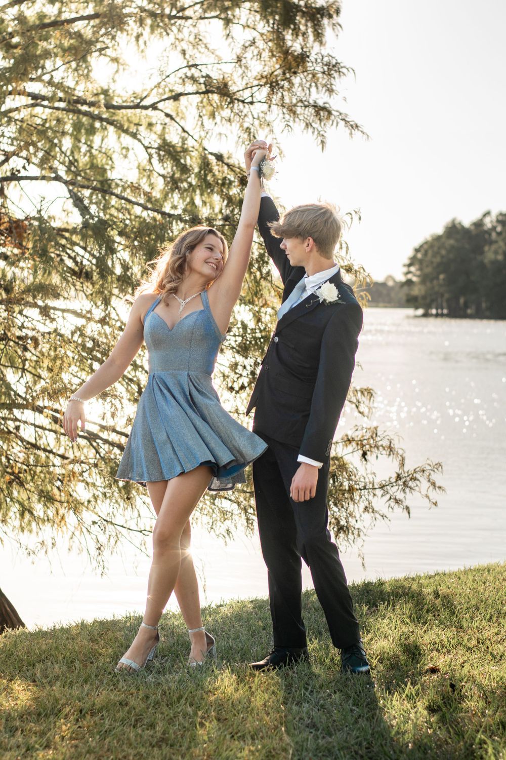 Dancing couple in formal wear twirl on grassy lakeside setting during golden hour.
